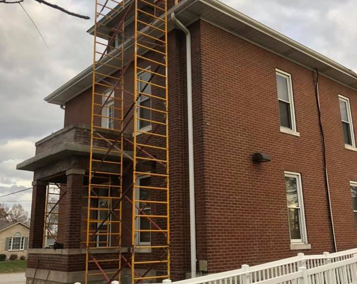 A red brick two-story house with white trim has scaffolding on the side. The sky is overcast, suggesting an ongoing construction or repair project.