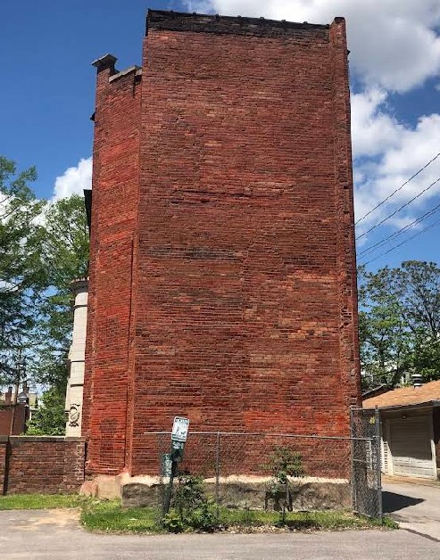A tall, flat, red brick building side stands against a blue sky with clouds. The scene is bordered by trees, a fence, and a paved path, creating an urban feel.