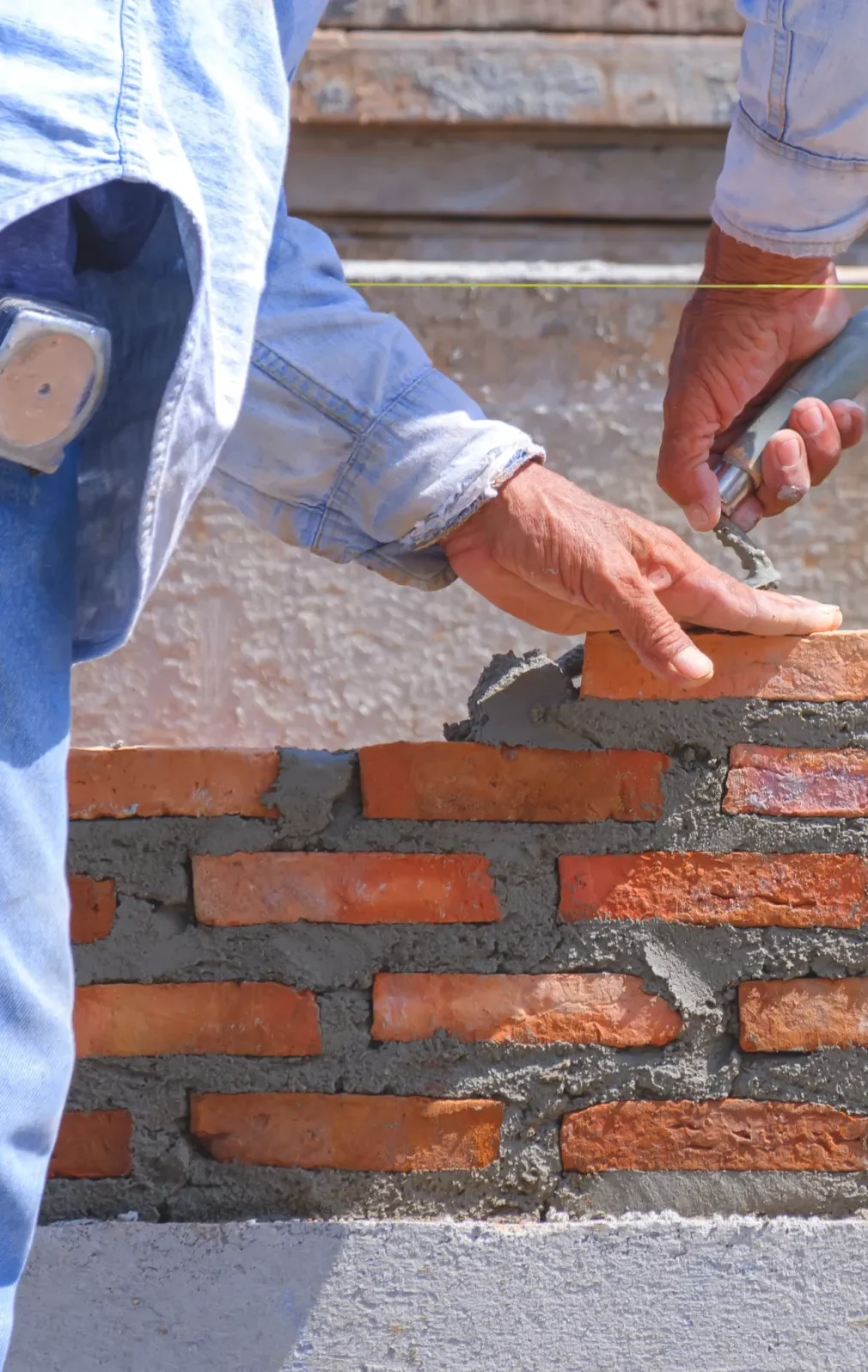 A worker in a denim shirt carefully lays bricks on a wall, smoothing mortar with a trowel. The scene conveys precision and craftsmanship in construction.