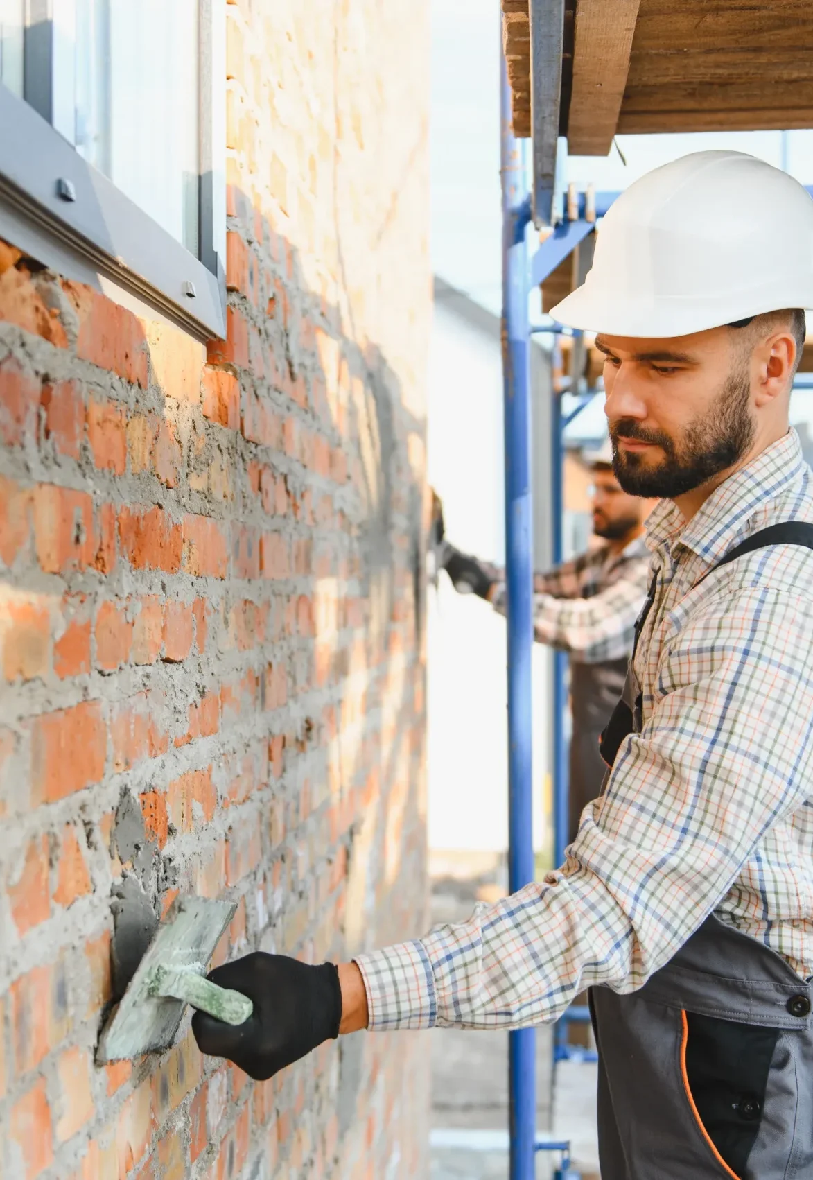 A construction worker in a white hard hat and plaid shirt applies cement to a brick wall, focused and intent. Sunlight casts shadows, creating a warm tone.