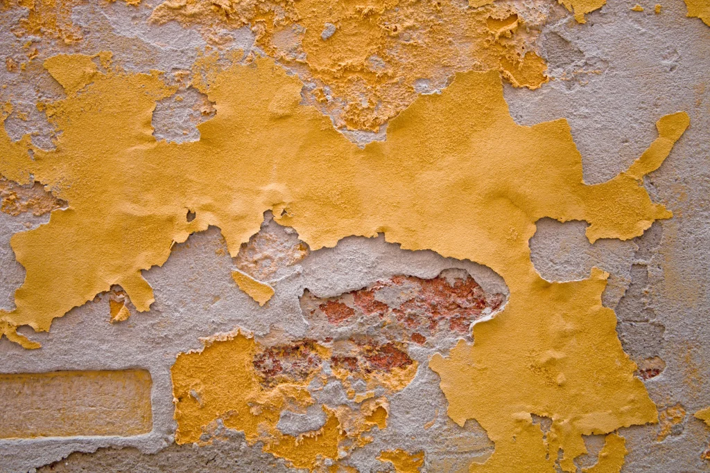 Close-up of a weathered wall with peeling yellow paint revealing textured red brick and gray plaster underneath. The surface looks aged and rustic.