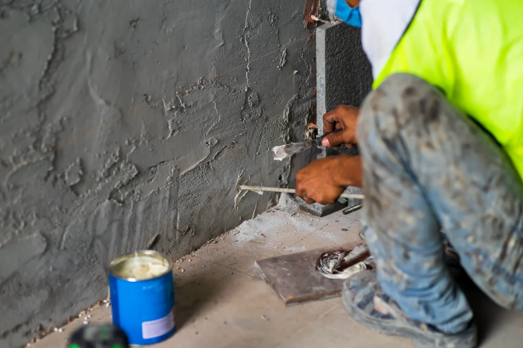 A construction worker in a neon vest and jeans applies putty to mortar on a wall. Nearby, an open blue can of putty and tools are visible on the floor.