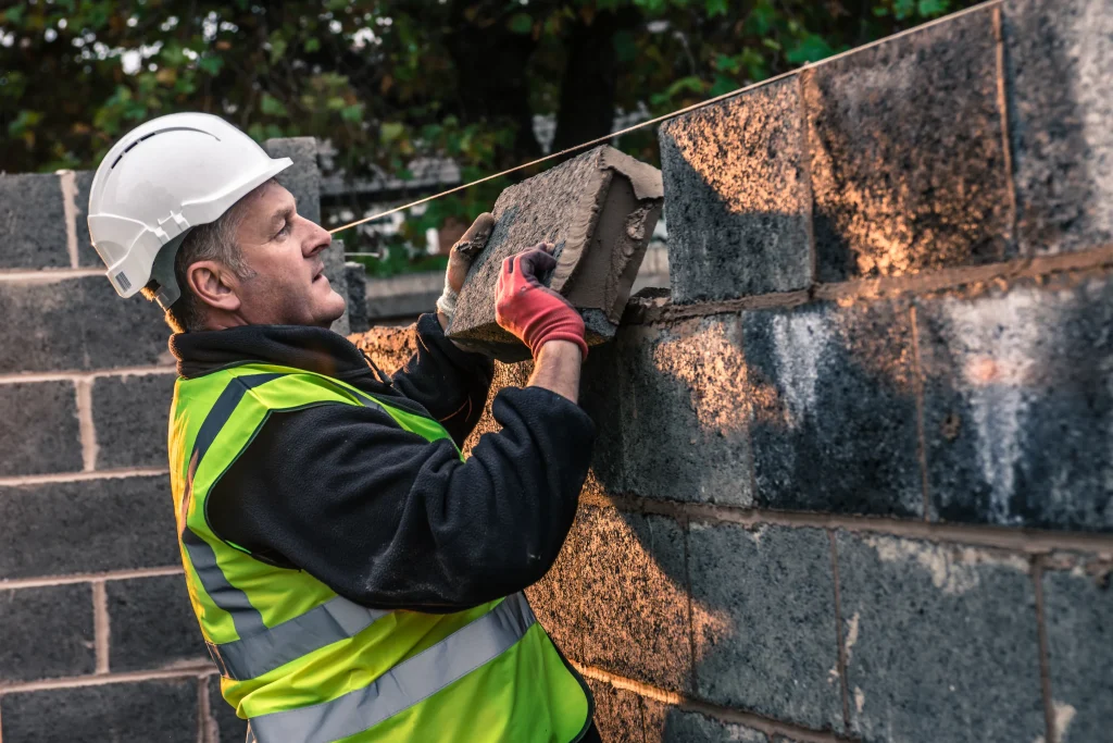A construction worker in a hard hat and high-visibility vest lays bricks on a wall at sunset. The scene conveys focus and craftsmanship.